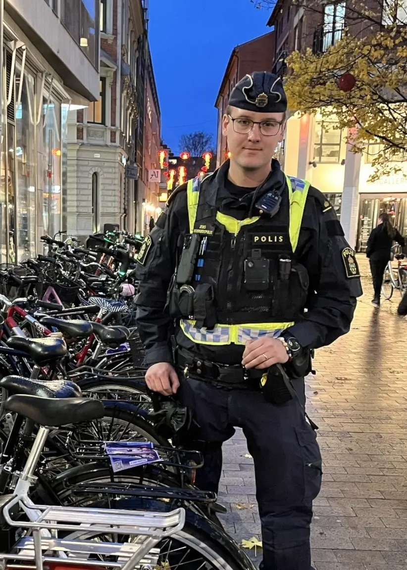 En polis står på en gata med cyklar parkerade längs trottoaren, omgiven av butiker och fotgängare under skymning.