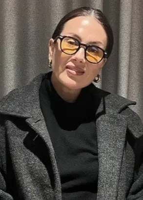 En person klädd i en mörk kappa och svart tröja står framför ett grått draperi och håller något i händerna.