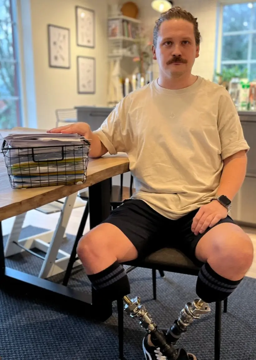 Alexander Helander från Ulricehamn sitter vid ett bord i ett modernt kök, klädd i t-shirt och shorts, med en bunt papper och en laptop framför sig. På bordet syns även flera dokument.