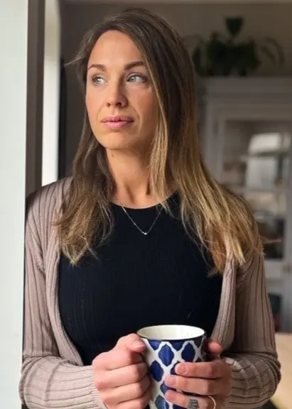 En kvinna står vid ett fönster inomhus och håller i en mönstrad kopp. Utanför syns en snötäckt trädgård och hus i bakgrunden. Inredningen är ljus med en vit skänk i rummet.