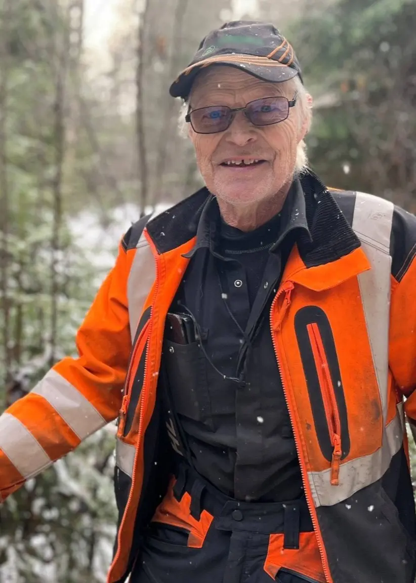 Skogsägaren Sven Olofsson i Hovmantorp i orange skyddsväst och mörka kläder står i en snöig skog och pekar på marken, omgiven av granar och vinterlandskap.