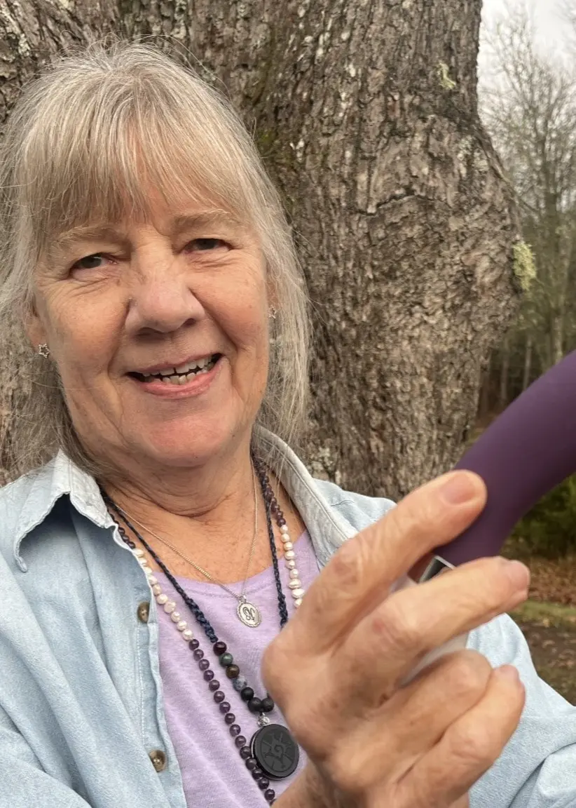 En person håller två föremål, ett träobjekt och ett lila föremål, framför en stor trädstam i en utomhusmiljö med gräsmatta och träd i bakgrunden.