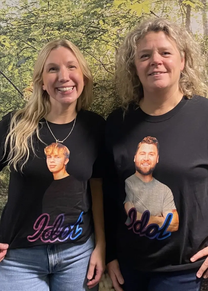 Karin Backlund och Stina Erixon poserar framför en vägg med en tapet med skog, båda klädda i svarta tröjor med bilder på sina söner som tävlar i musiktävlingen Idol.