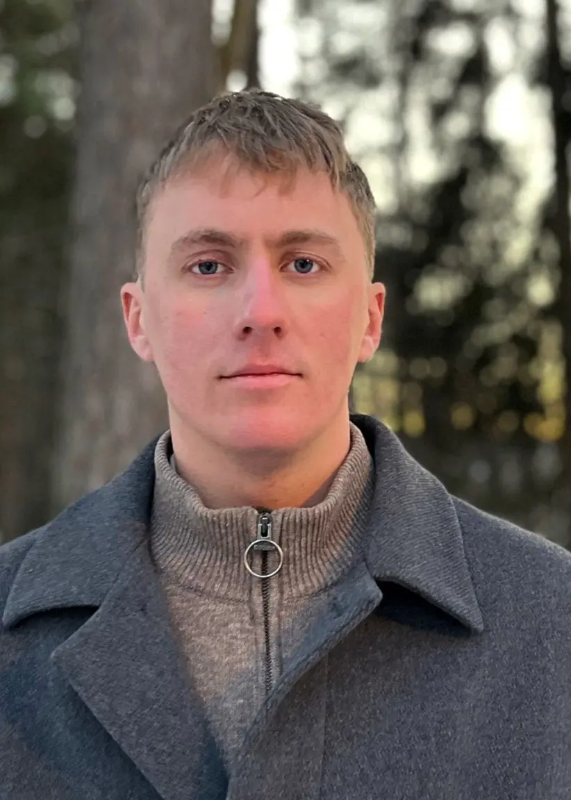 Gustav Lidenholm, officer vid regementet i Enköping, står utomhus klädd i en grå kappa med en vinterlandskap i bakgrunden där solen går ner bakom snötäckta träd.