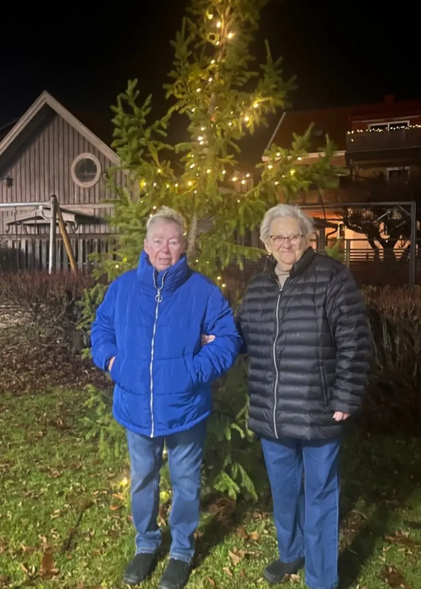 Marie-Louise Johansson och Maja Davidsson står vid en upplyst gran på Biblioteksgatan i Växjö under kvällstid. Granen är dekorerad med ljusslingor och omgivningen är mörk.