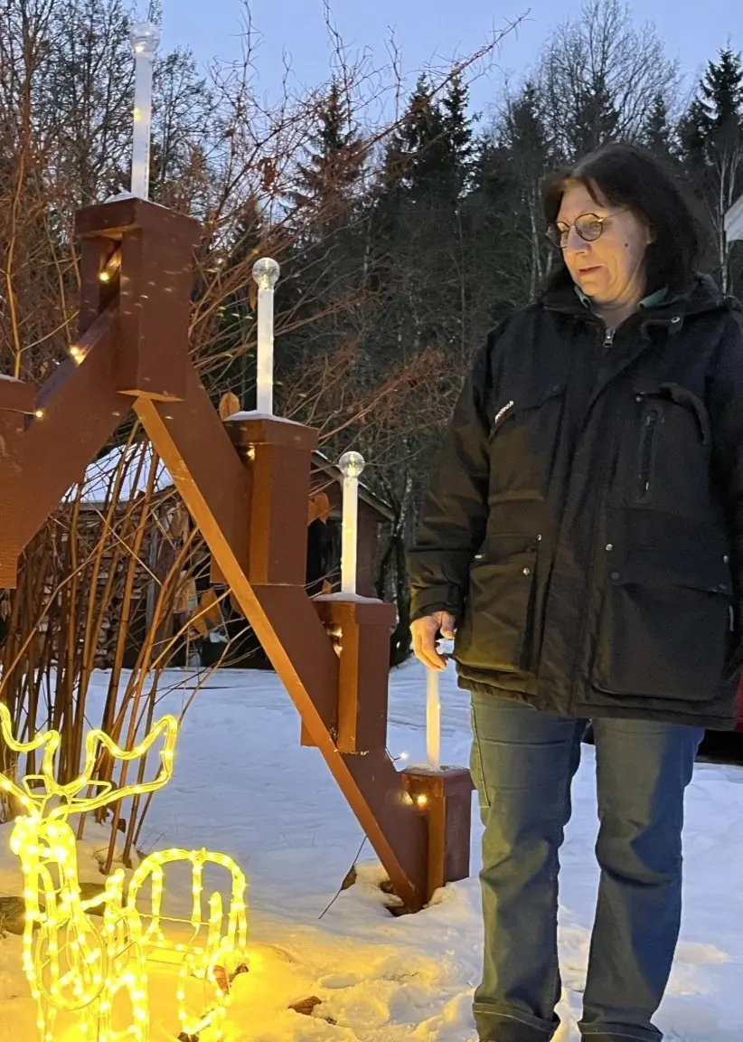Inger Dagh står utomhus vid en snötäckt trappa dekorerad med ljusstakar och en lysande ren i julbelysning. En röd bil syns i bakgrunden vid ett hus.