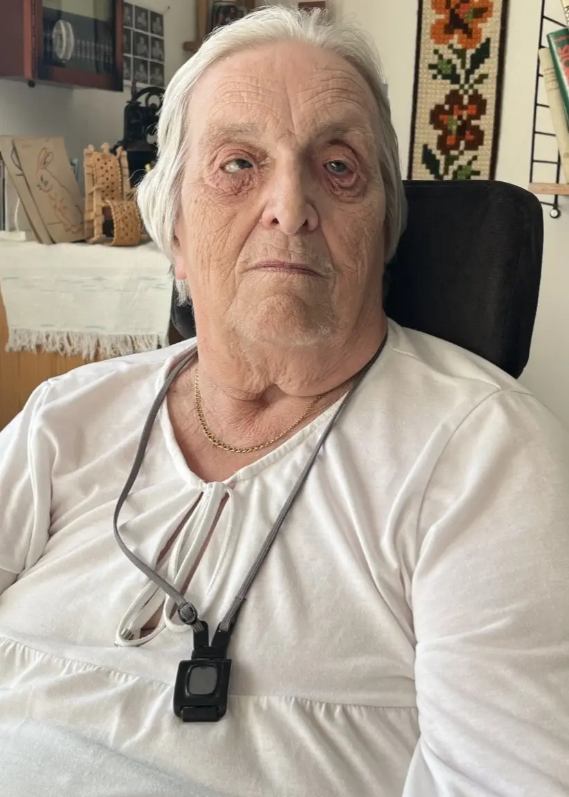 Irene sitter i en stol i sitt hem i Östhammars kommun. Hon har en vit tröja och ett trygghetslarm runt halsen. Bakgrunden visar hyllor med böcker och flera fotografier på väggen.