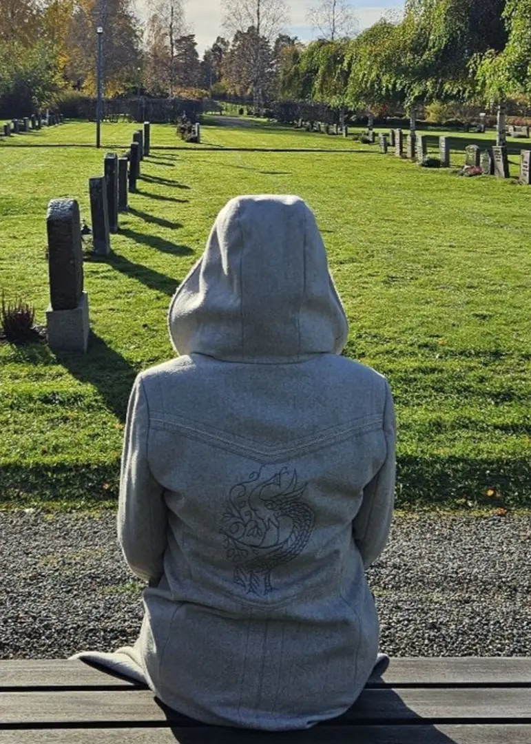 En person i grå jacka sitter på en bänk vid en kyrkogård under en solig dag. Gravstenar syns i bakgrunden, omgivna av grönska och höstfärger.