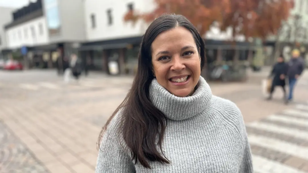 Cecilia Soto Thompson står utomhus på en stadsgata med höstfärgade träd i bakgrunden. Hon bär en grå stickad tröja och omgivningen inkluderar byggnader och människor som promenerar.