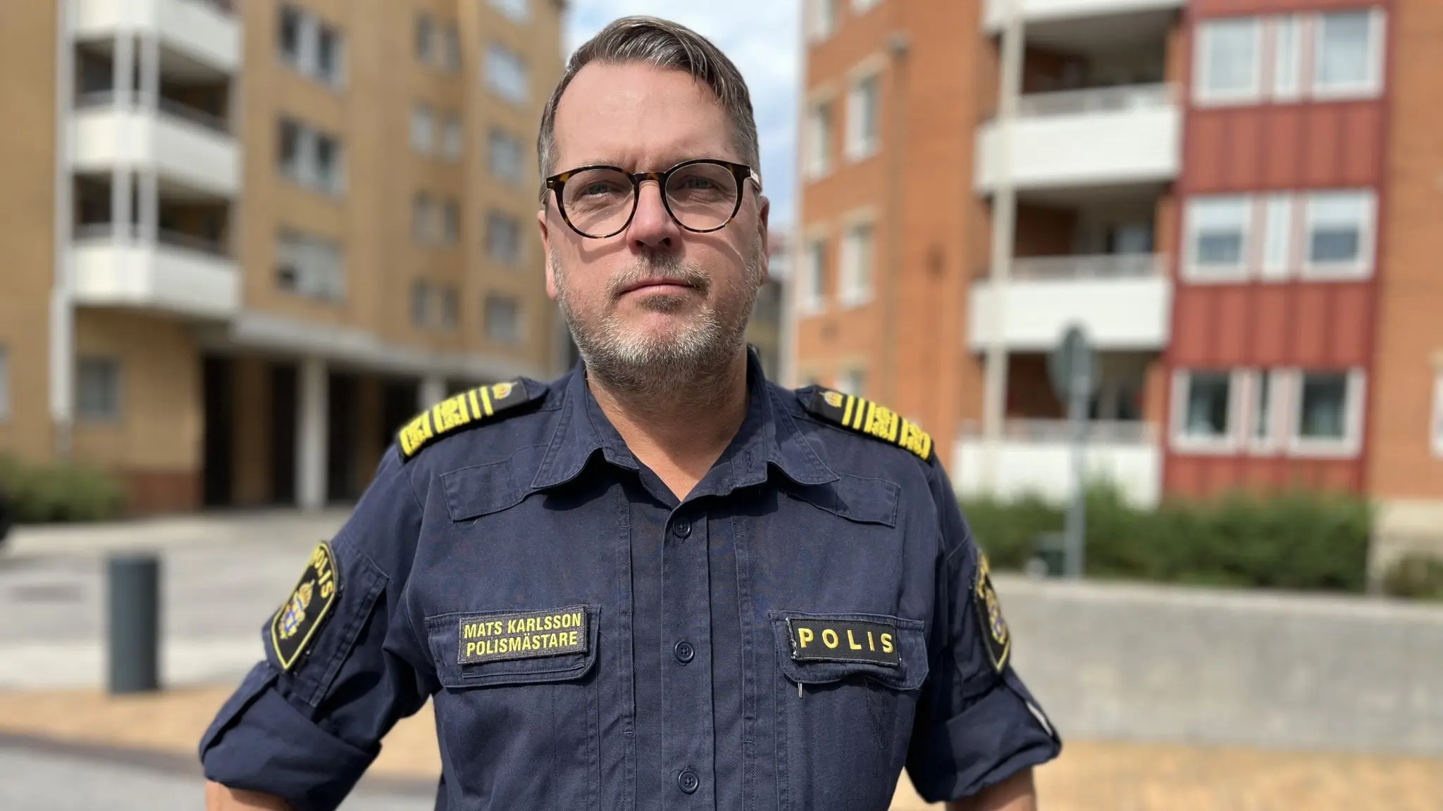 Mats Karlsson, klädd i en polisuniform, står framför flerfamiljshus i ett bostadsområde.