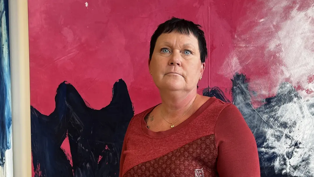 Ilho Lindström står framför en färgstark vägg med rosa och blå abstrakta målningar, iklädd en röd tröja med mönster. Bakgrunden ger en konstnärlig känsla.