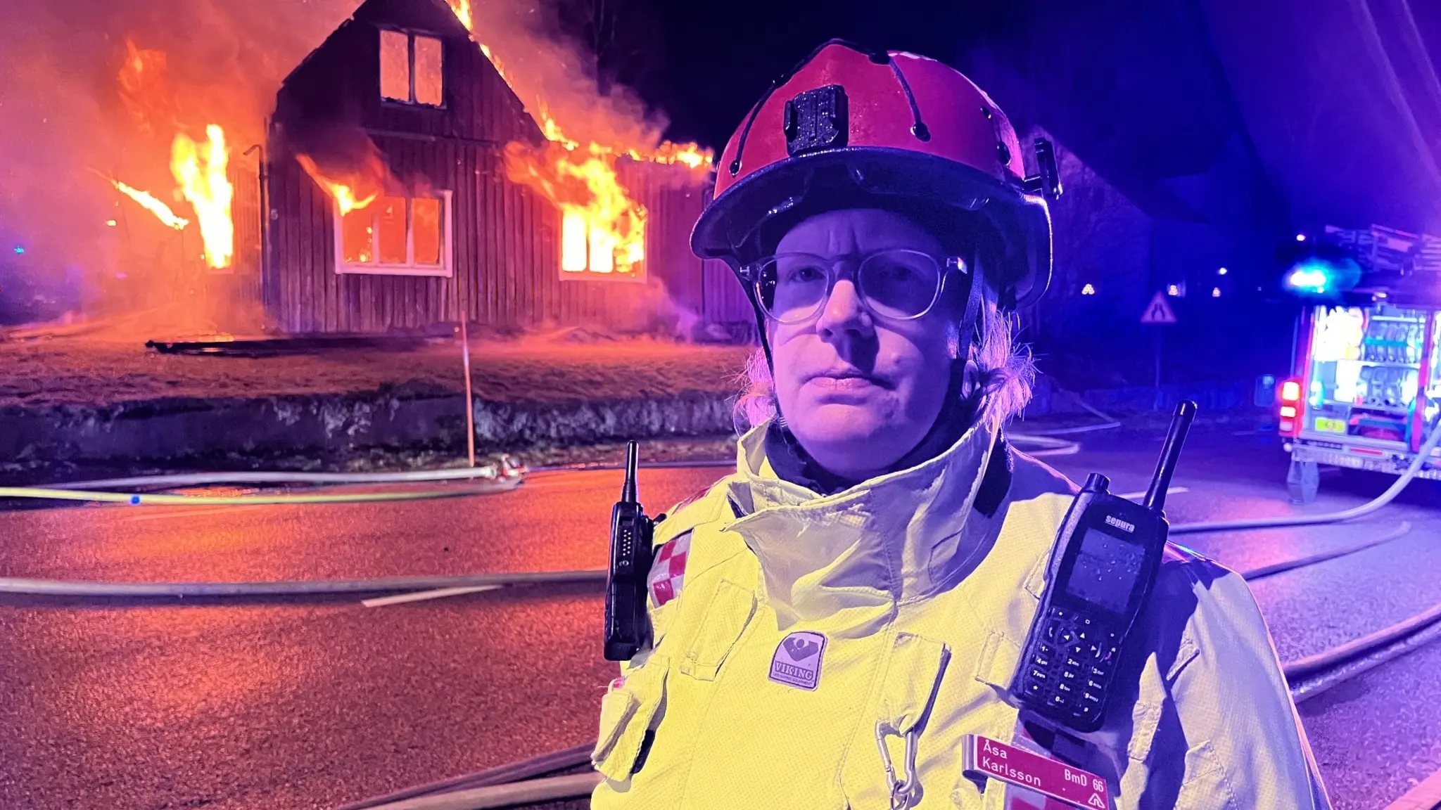 En brandman i gul skyddsutrustning och röd hjälm står framför ett brinnande trähus på natten. Eldslågor syns tydligt från byggnaden och räddningstjänstens fordon är i bakgrunden.