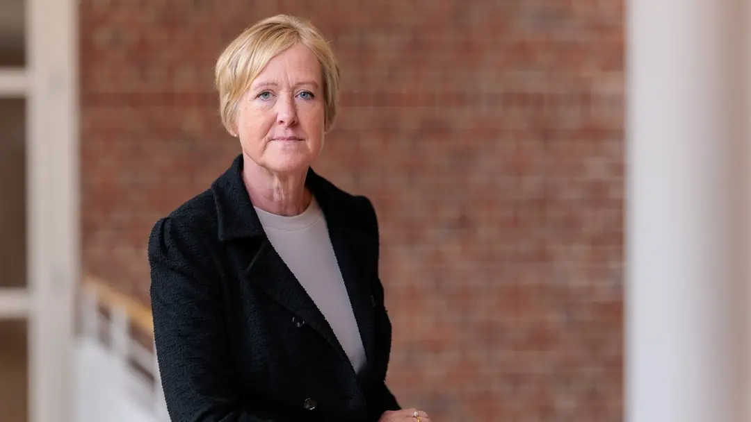 Ewa Ekman, klädd i mörk kavaj och vit topp, står vid ett räcke inomhus med en tegelvägg i bakgrunden. Bilden är tagen i en ljus miljö med fokus på en professionell framtoning.