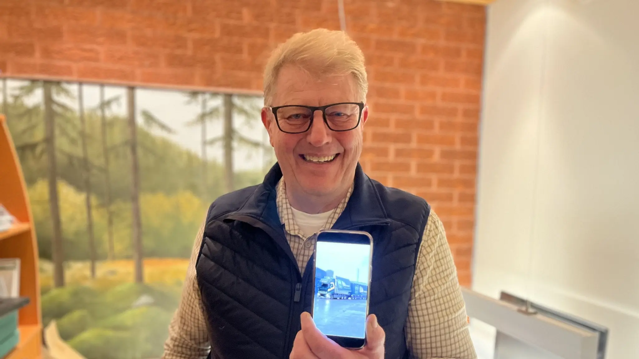 En person håller upp en smartphone som visar en bildskärm. Bakgrunden består av en tavla med naturmotiv och en vägg av rött tegel. Personen bär en stickad tröja och en mörk väst.