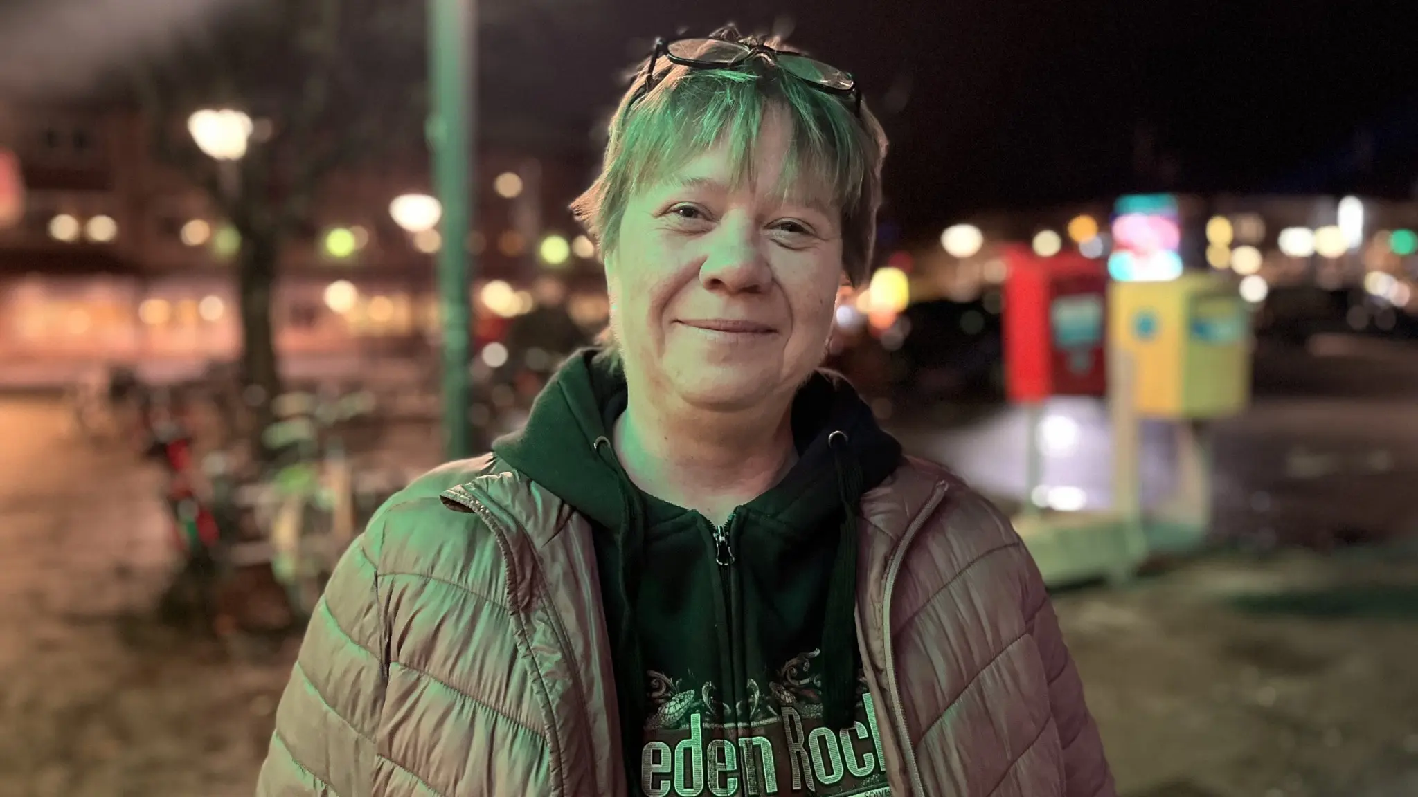 Maria står utomhus på en upplyst kvällsbild med en ljus jacka och mörk hoodie. Bakgrunden visar gatubelysning och en offentlig miljö.