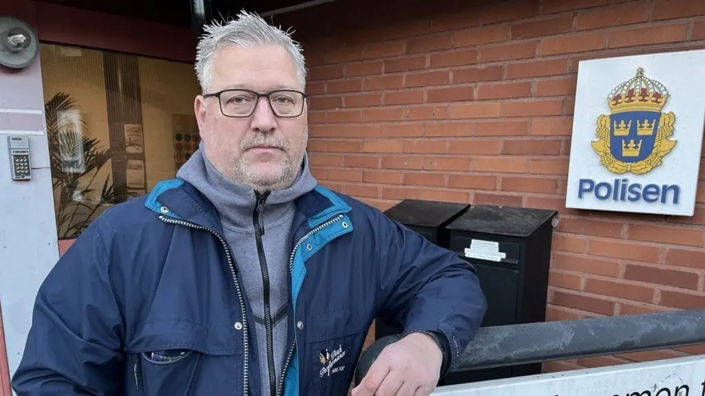 Hans Sjöberg, kommunpolis i Ljungby, står utomhus vid en tegelvägg med en skylt där det står 'Polisen'. Han bär en blå jacka och lutar sig mot ett staket.