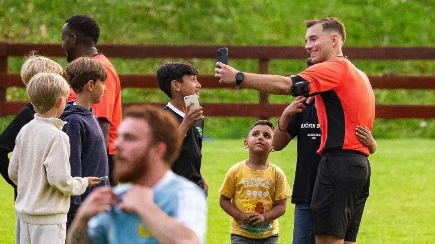 En man i fotbollsdomartröja, identifierad som Taib Sisic, talar med en grupp barn på en gräsplan. Barnen är klädda i fritidskläder och verkar delta i en aktivitet utomhus.