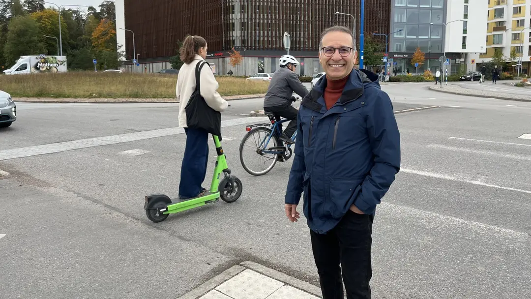 En person står vid en trafikkorsning, medan en annan kör en elsparkcykel och ytterligare en cyklar. En modern byggnad och andra stadsmiljöer syns i bakgrunden.