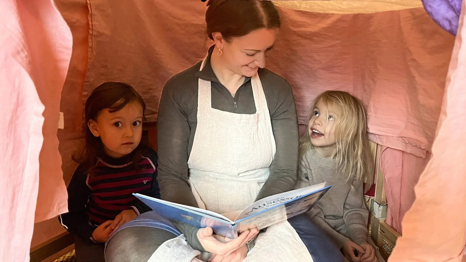 Siri Lind, klädd i vit förkläde, sitter inne i ett tältliknande arrangemang och läser en bok för två barn. Tältet är gjort av färgglada tyger och skapar en mysig atmosfär.