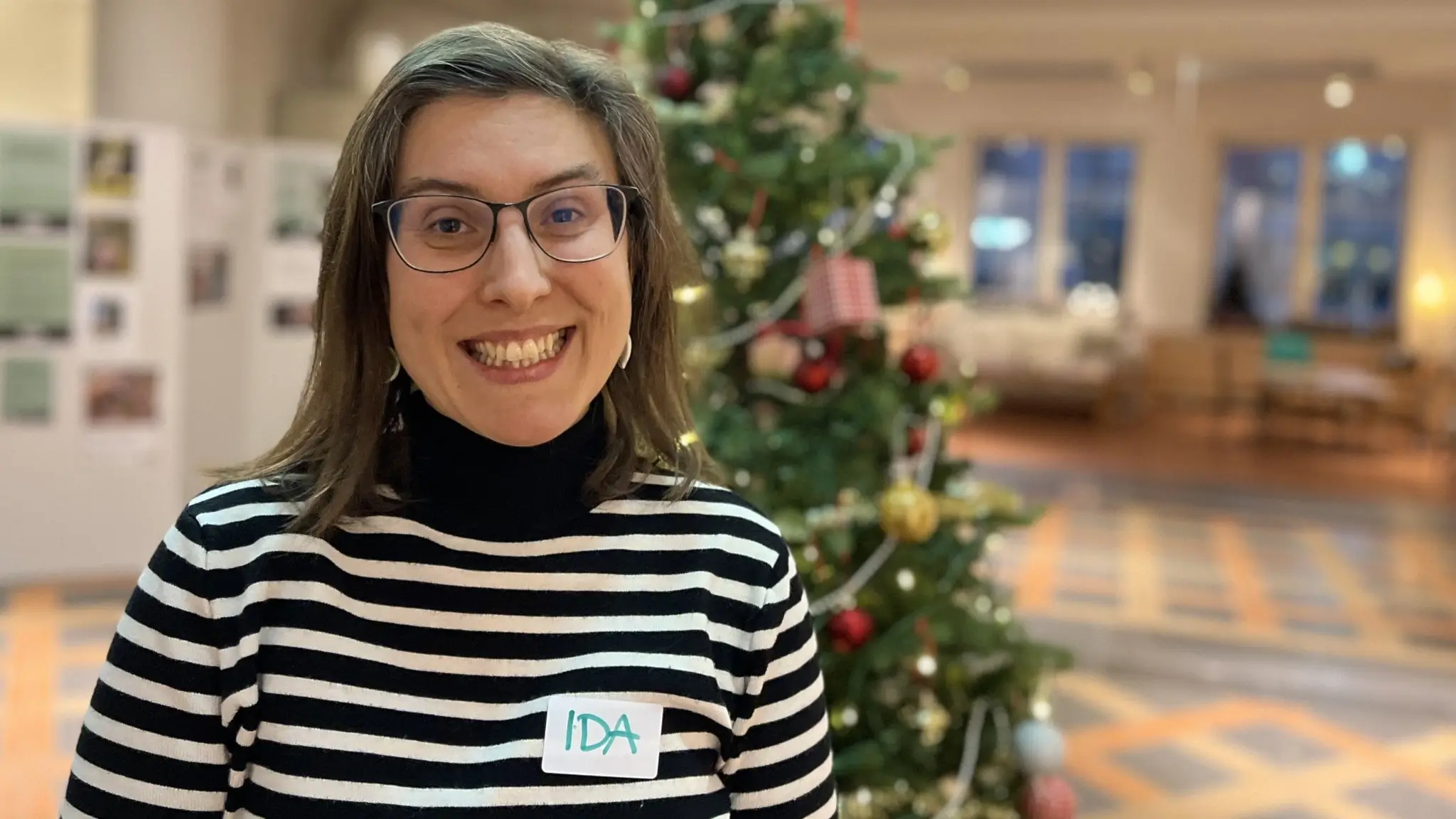 Ida Lindhagen, projektledare för Uppsala stadsmissions julklappsbutik, står framför en pyntad julgran i en ljus inomhusmiljö.