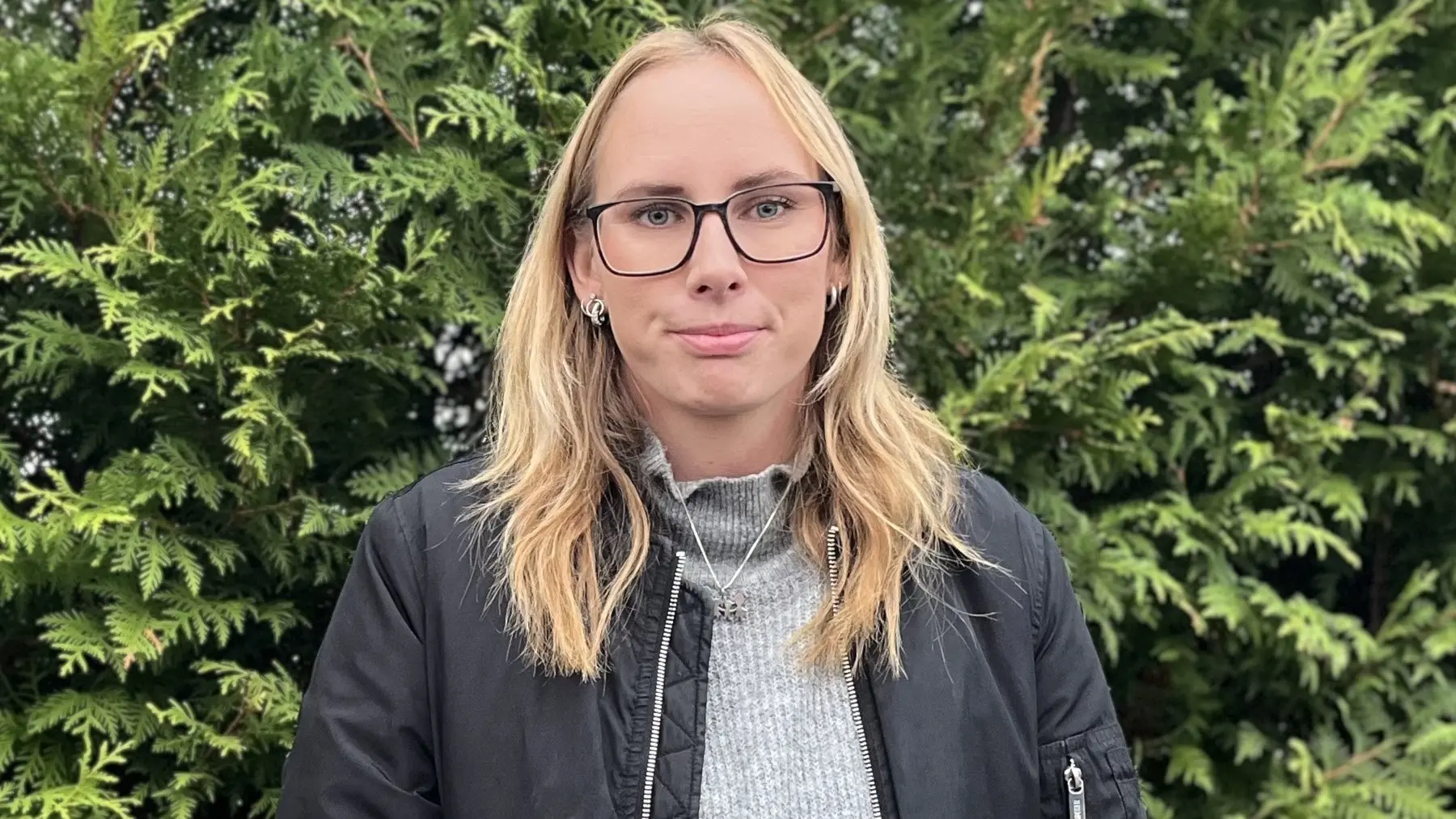En kvinna med långt, blont hår och glasögon står framför en grön häck iklädd en svart jacka och ljusgrå tröja.