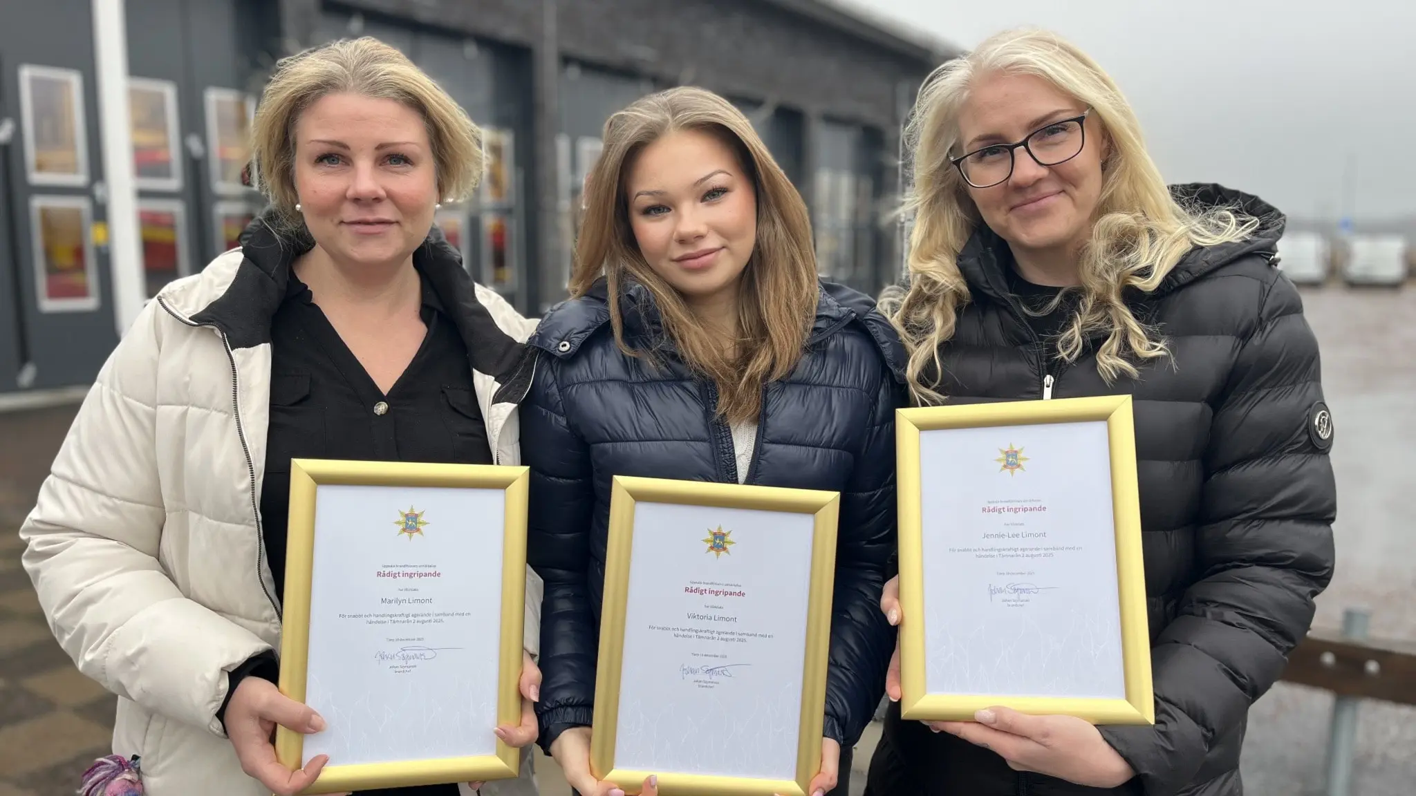 Jennie-Lee, Marilyn och Viktoriastår utomhus och håller diplom med guldram framför en byggnad. De är klädda i jackor och ser ut att ha deltagit i en ceremoni eller mottagit utmärkelser.