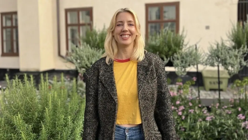 Veronica Maggio står utomhus framför en byggnad med stora fönster, omgiven av grönskande växter. Hon bär en gul tröja, en mörk kofta och jeans, med avslappnad kroppshållning.