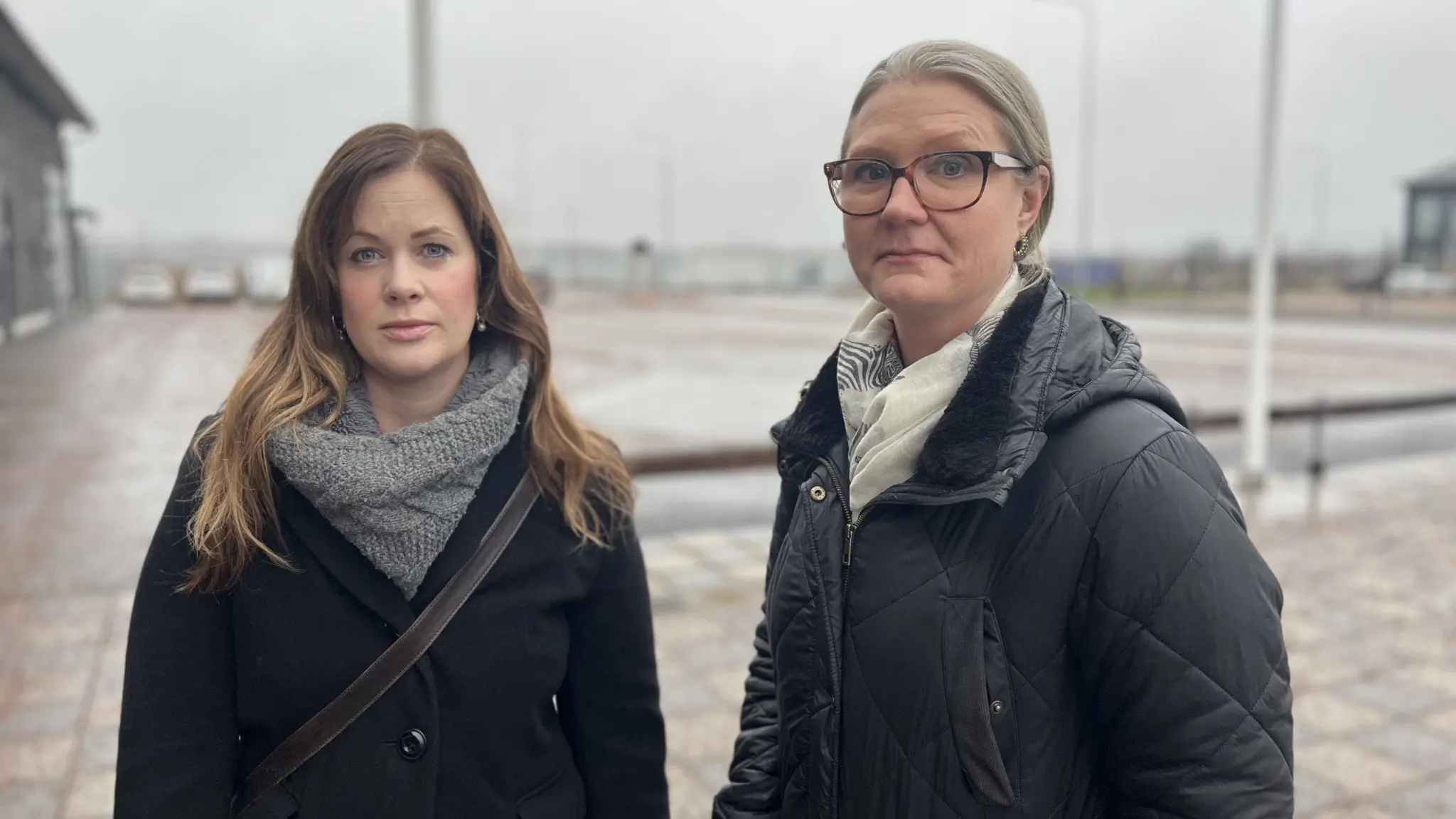Viktoria Söderling och Sara Sjödal står utomhus på en gråmulen dag vid en skolgård med asfalt och bänkar i bakgrunden. De bär mörka jackor och halsdukar, och området är omgärdat av öppna ytor.
