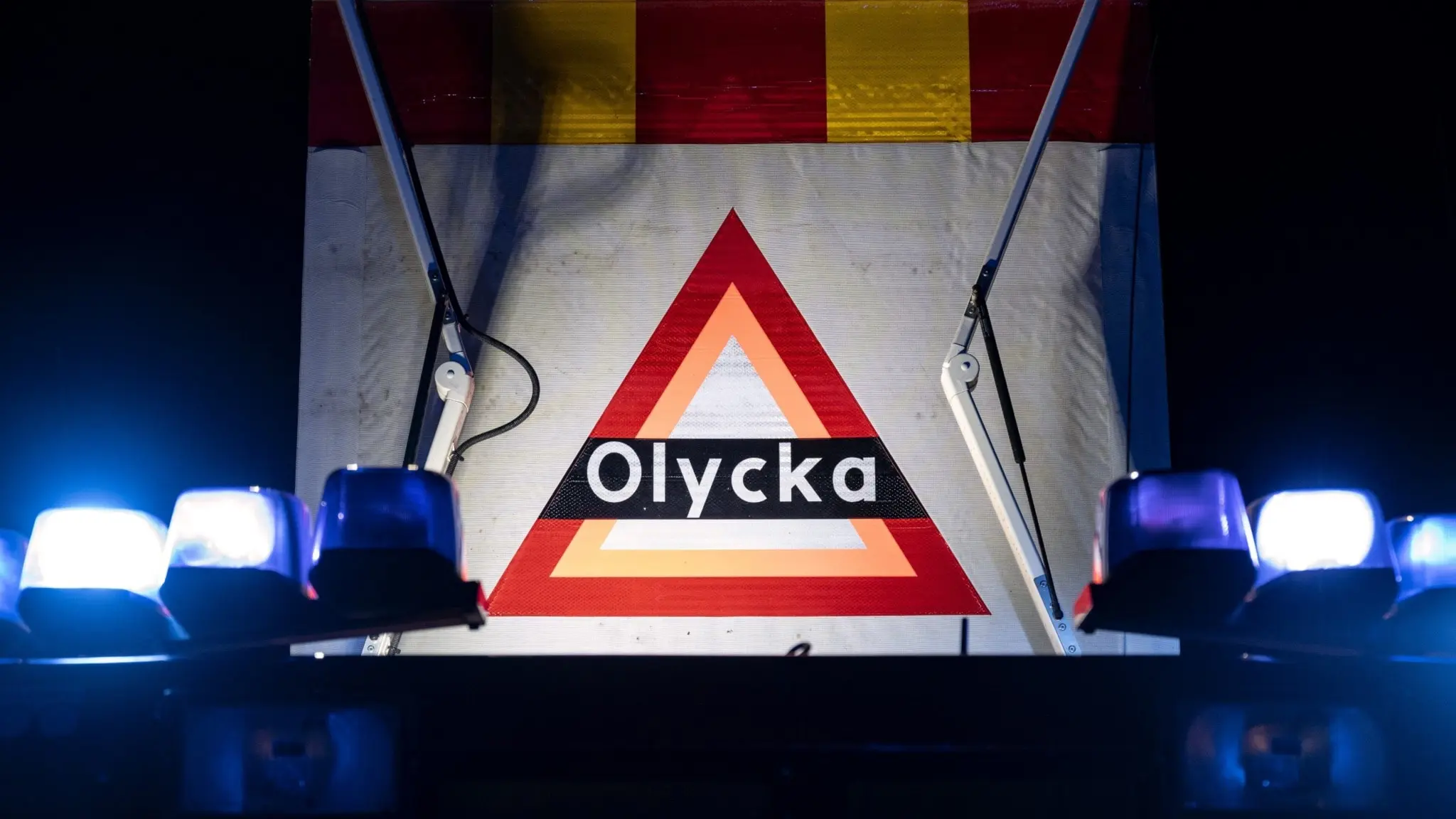 En varningsskylt med texten 'Olycka' är upplyst av blåljus från räddningstjänsten vid en olycksplats. Skylten markerar närvaro av räddningspersonal.