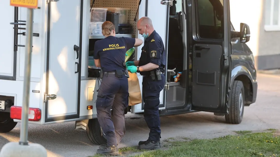 Poliser undersöker bevismaterial vid en öppnad polisbil, klädda i uniform och skyddshandskar. Händelsen sker på en gata med grönska i bakgrunden, vilket tyder på en pågående utredning.