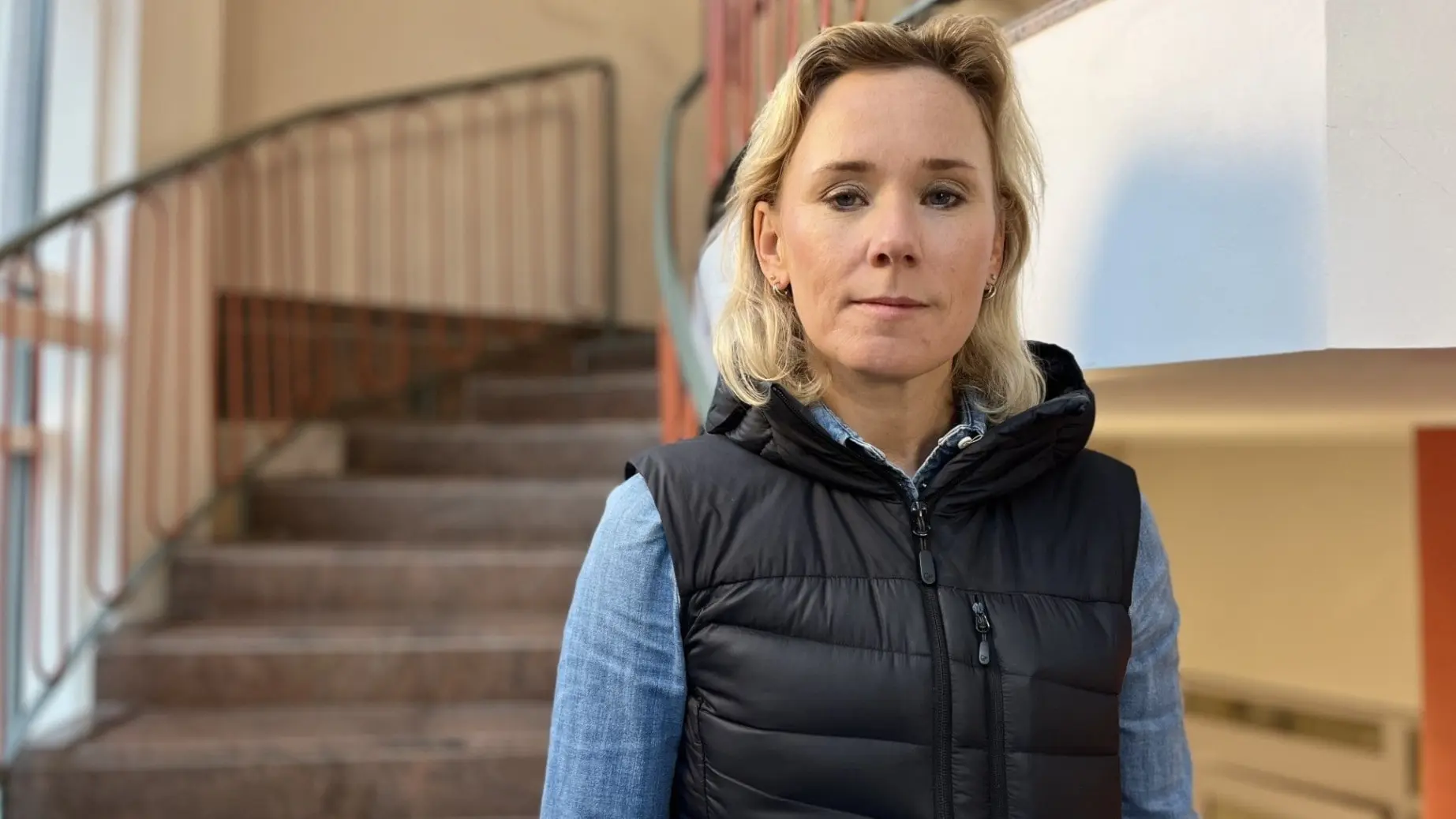 En person i blå skjorta och svart väst står framför en trappa inomhus. Bakgrunden visar en ljus vägg och ett räcke av metall.