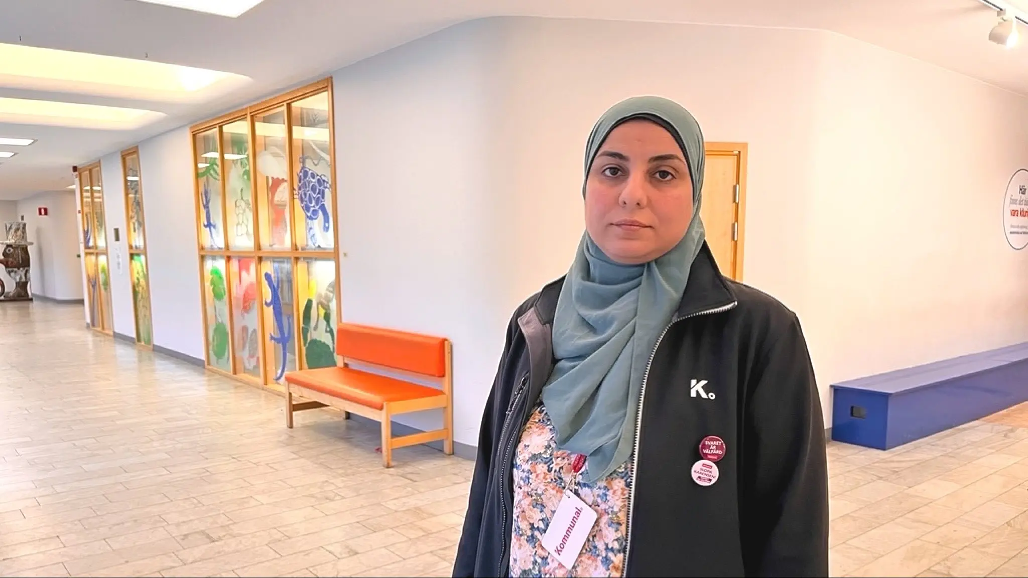Sumaia Eizuldeen, sektionsordförande i Kommunal, står inomhus. Bakgrunden visar en ljus korridor med färgglada väggdekorationer och en orange bänk.