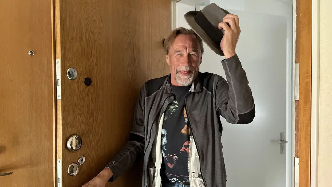 Timo, tidigare hemlös, står vid dörren till sin nya lägenhet i Uppsala och lyfter på hatten. Han lyfter på hatten som att hälsa och håller på att öppna dörren med ena handen.