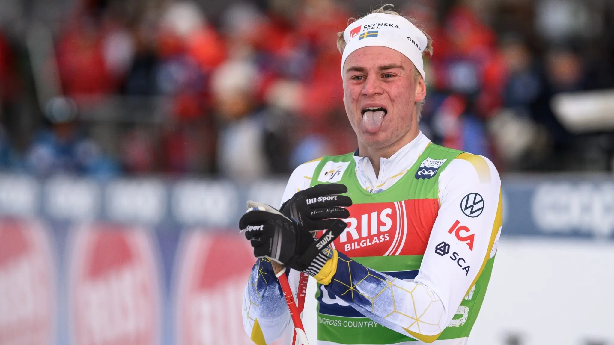 En längdskidåkare i tävlingsdräkt med grönt västskydd deltar i en skidtävling. Bakgrunden visar publik i röda kläder och ett tävlingsområde med reklamskyltar.