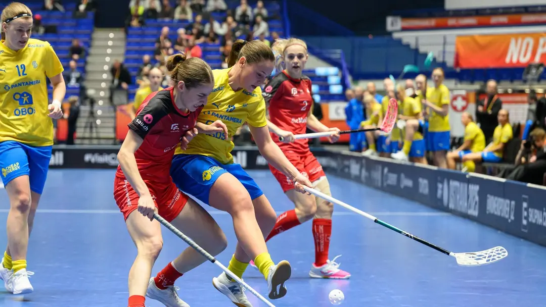 Svenska innebandylandslaget i gula tröjor möter motståndare i röda tröjor i en intensiv kamp om bollen under en match på blått golv, med publik i bakgrunden.