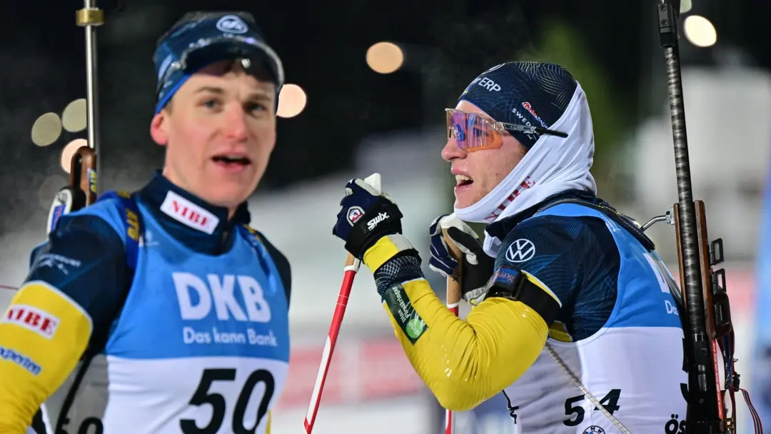 Två manliga skidskyttar i tävlingsdräkter med gevär på ryggen står på en belyst arena under en tävling. De interagerar med varandra, och sponsorskyltar syns i bakgrunden.