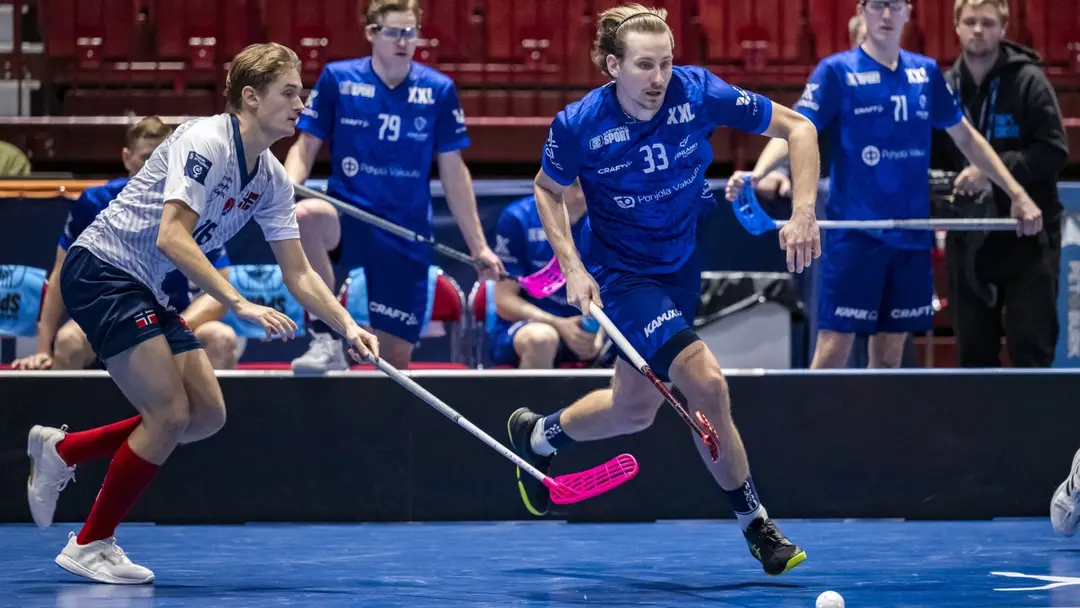 Två innebandyspelare i intensiv kamp om bollen, en i blå tröja och en i vit tröja, på en inomhusbana med åskådare och avbytarbänk i bakgrunden.