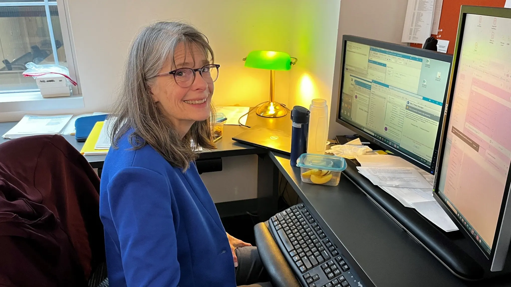Kvinna framför två dataskärmar vid skrivbordet tittar upp. Har glasögon och i blå kavaj.