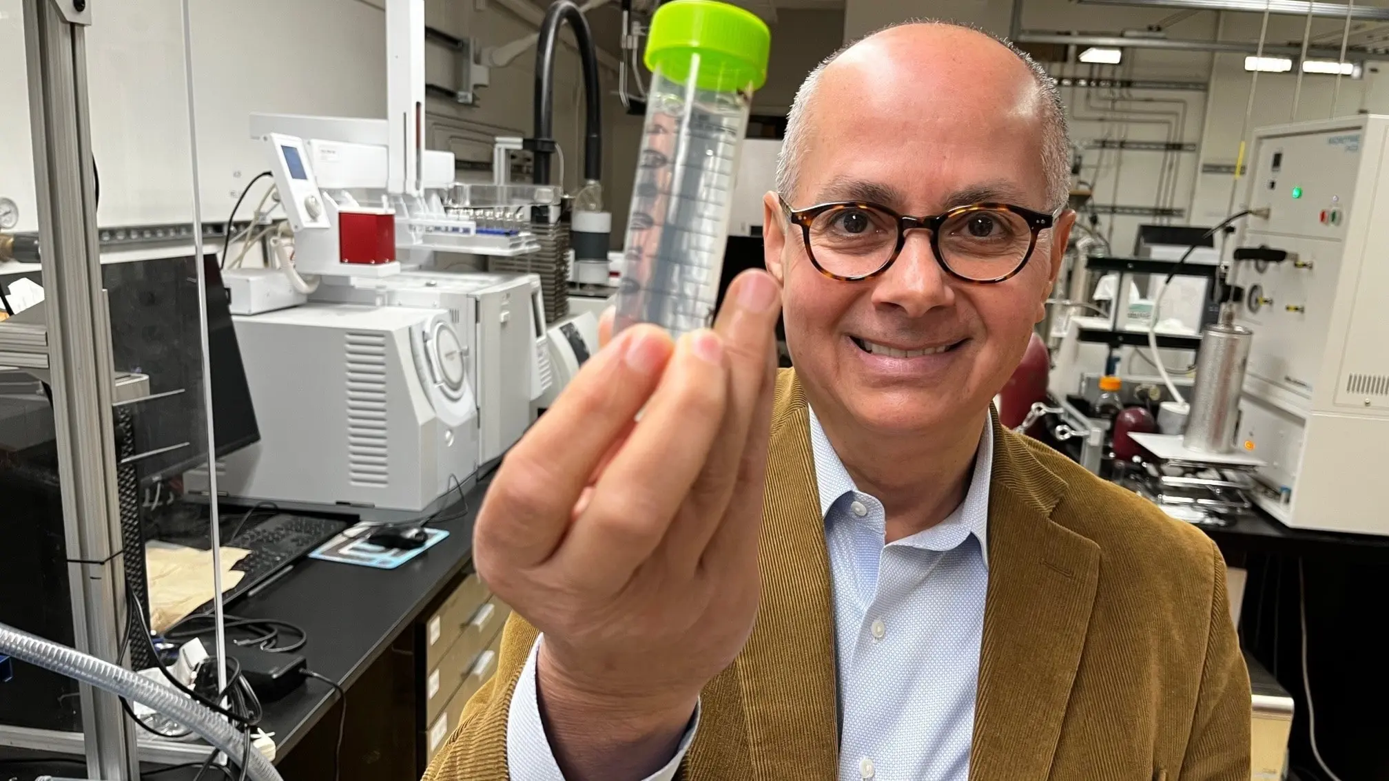 Nobelpristagaren Omar Yaghi med ett provrör med grönt lock som innehåller en lösning med ett metall-organiska ramverk. I bakgrunden flera maskiner i labbmiljö.