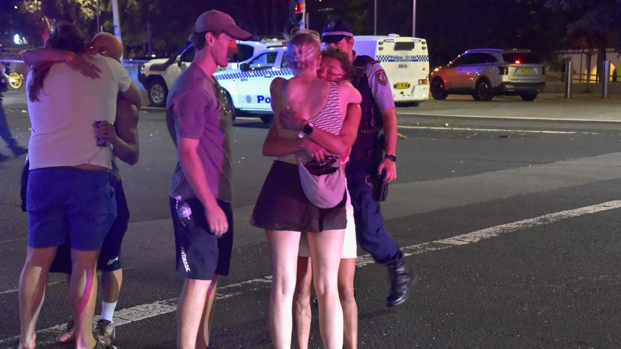 Människor samlas och visar känslor efter en skjutning vid Bondi Beach i Sydney. Polisfordon och uniformerade poliser syns i bakgrunden, tillsammans med personer som kramar varandra i chock och sorg.