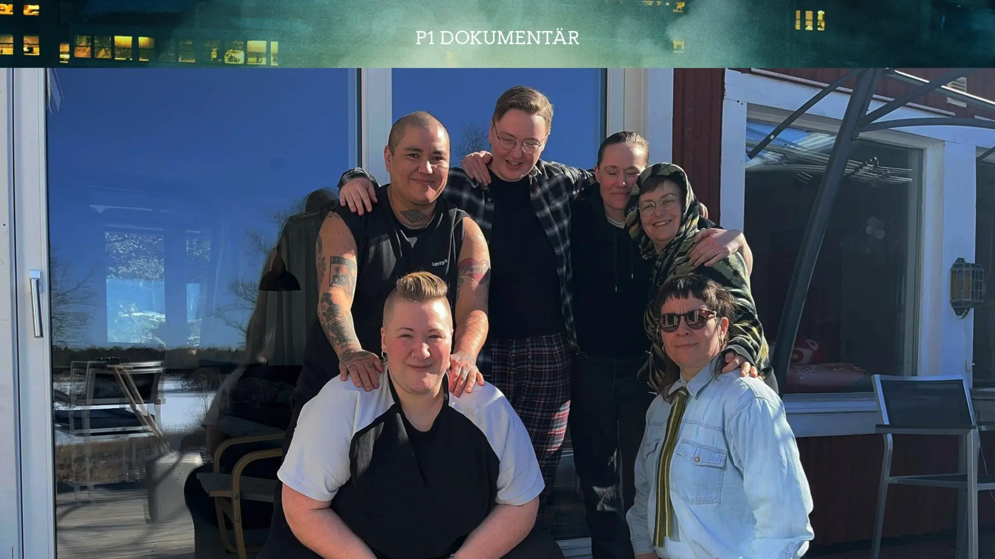 6 personer som står framför en stuga i solen. Två av dem sitter på knä framför fyra av dem som står i rad.