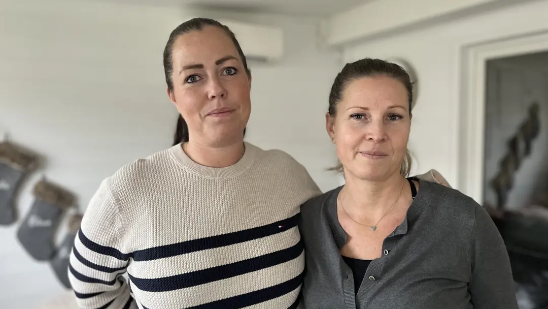 Två personer står nära varandra inomhus, den ena klädd i en vit tröja med svarta ränder och den andra i en grå kofta. I bakgrunden syns en ljus vägg.