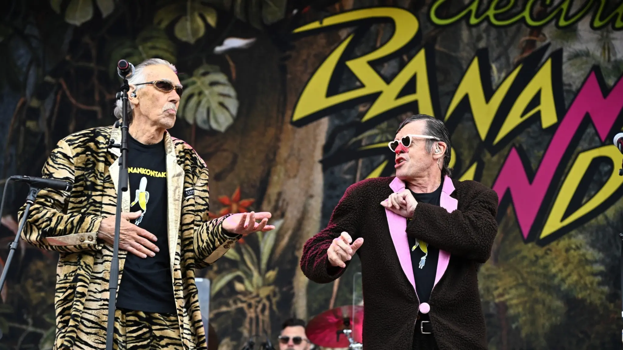 Electric Banana Band uppträder på Gröna Lund i Stockholm. Lasse Åberg i tigerkostym och Klasse "Banarne" Möllberg i sin bruna apkavaj står på scen med färgglad bandlogga i bakgrunden. En trummis syns bakom dem.