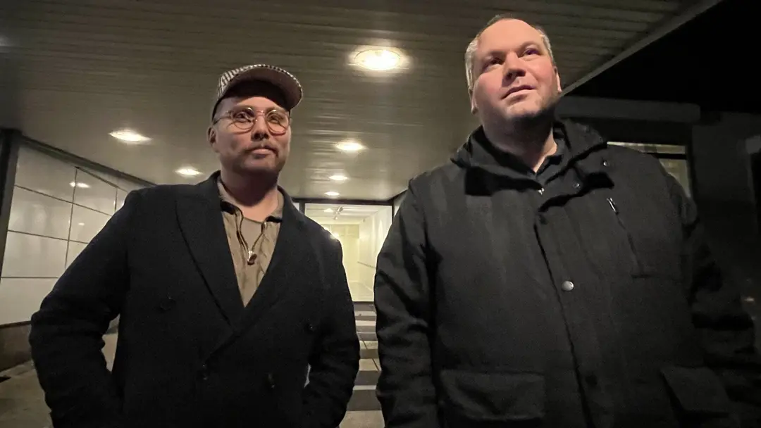 Två män står utomhus under ett tak med belysning. Den ena bär en keps och kappa, den andra en jacka. De befinner sig vid en byggnad med glasdörrar i bakgrunden.