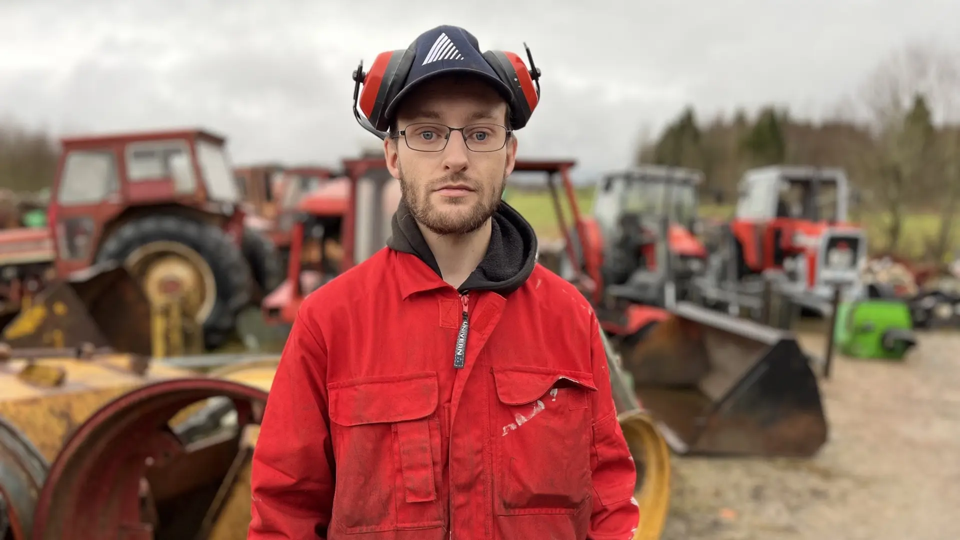 En person i röd arbetsoverall och hörselskydd står framför flera jordbruksmaskiner, bland annat traktorer och grävskopor, på en gårdsplan med gräs och träd i bakgrunden.