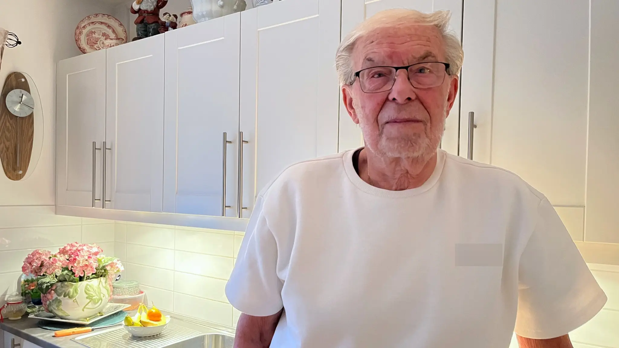 Johnny Claesson står i ett ljust kök med vita skåpluckor och en bukett blommor på köksbänken. En klocka och tallrikar syns på väggen i bakgrunden.