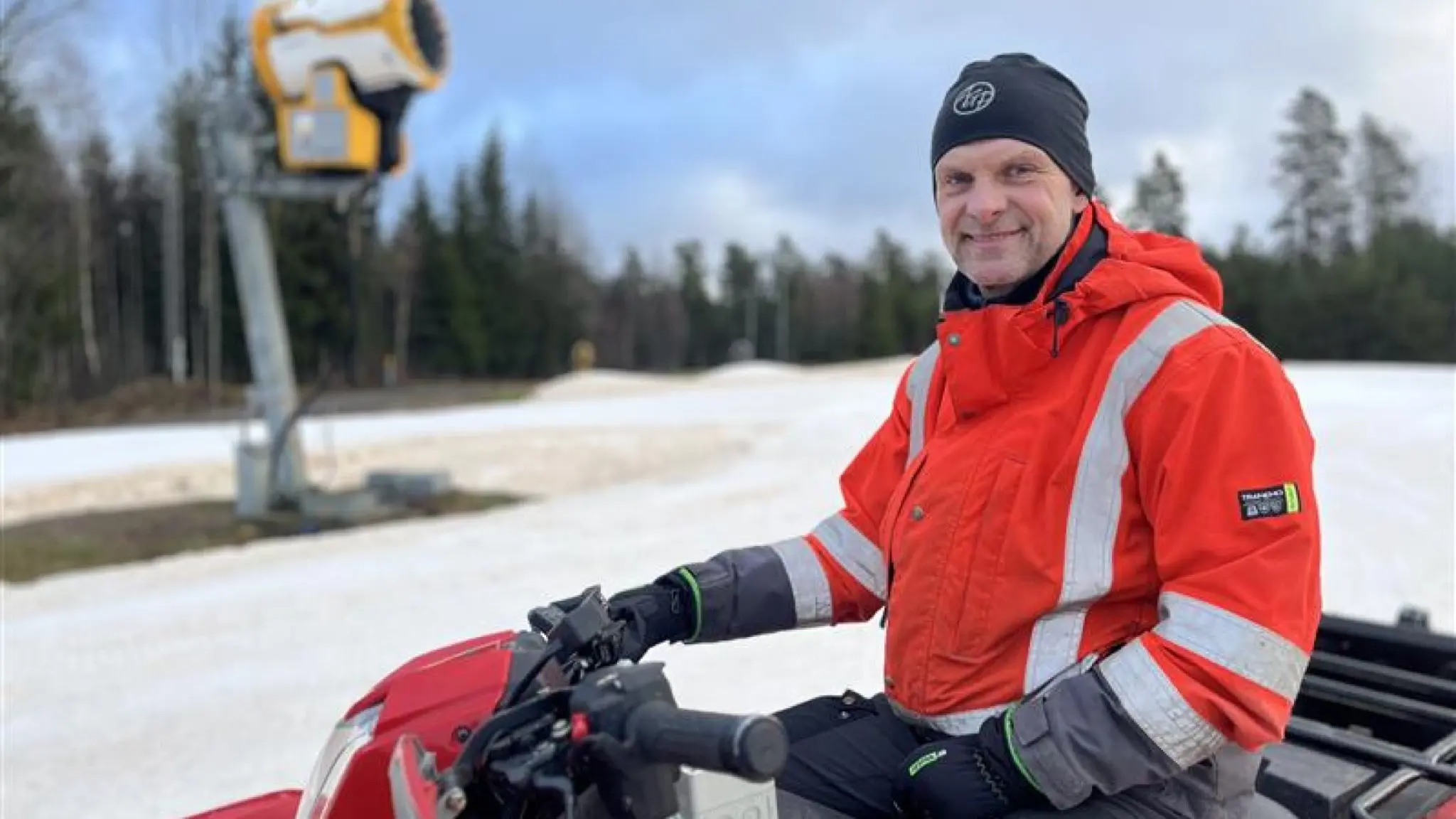 Kenneth Johansson sitter på en röd snöskoter klädd i en röd reflexjacka och mössa. Han befinner sig vid en snöbeklädd skidbacke omgiven av skog och en snökanon syns i bakgrunden.