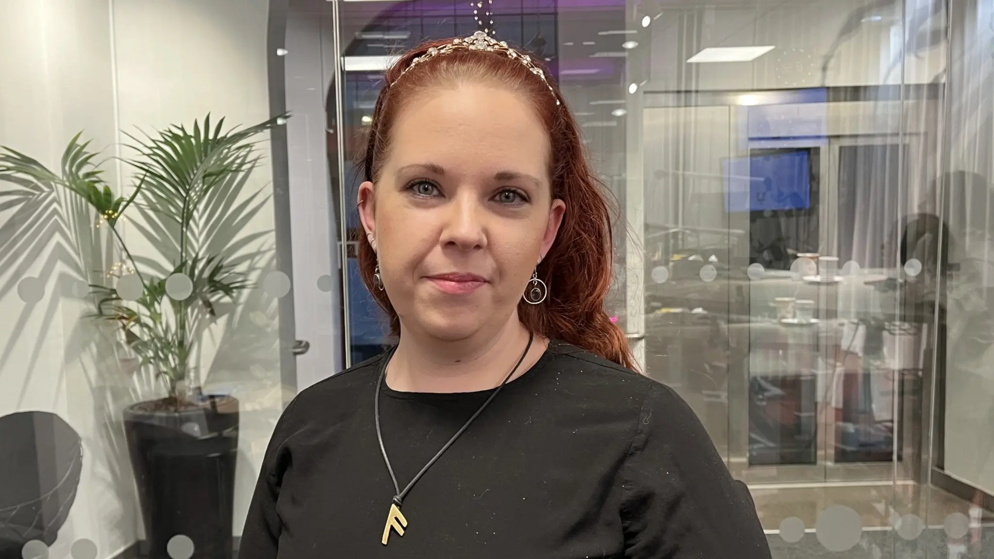 Emma Magnusson, nykter alkoholist, står inomhus i en modern kontorsmiljö med glasväggar och en grön växt i bakgrunden. Hon bär en svart tröja och ett halsband.