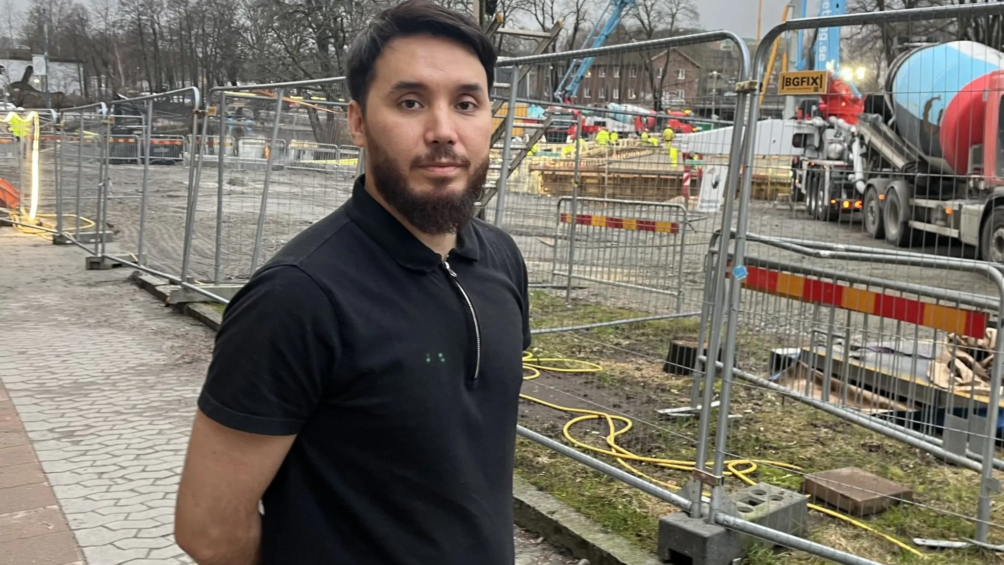Amin Hashemi, ägare av en restaurang i Borås, står framför ett byggarbetsområde med stängsel, byggmaskiner och grå himmel i bakgrunden.