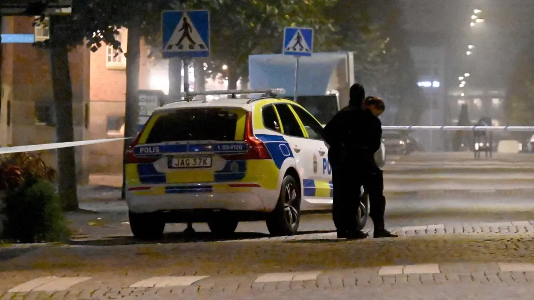 Polisbil och avspärrningsband på en mörk gata efter ett våldsbrott där en 17-årig flicka skadats allvarligt av knivhugg. Två poliser står vid bilen, området är tomt och upplyst av gatlampor.